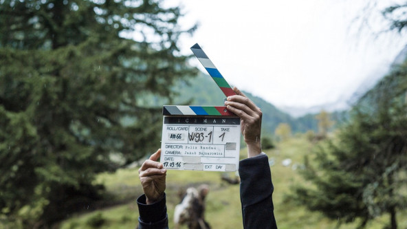 A film clapperboard can be seen in the middle of the picture. Behind it is a movie scene in a rainy forest.
